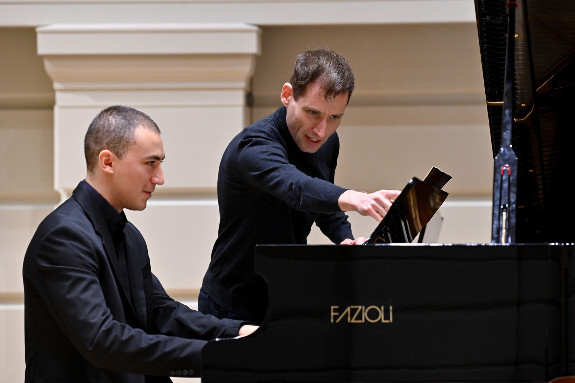 A man wearing a black shirt, pointing at the music sheet on the piano stand, with a younger man wearing smart black shirt, playing on the piano on stage.